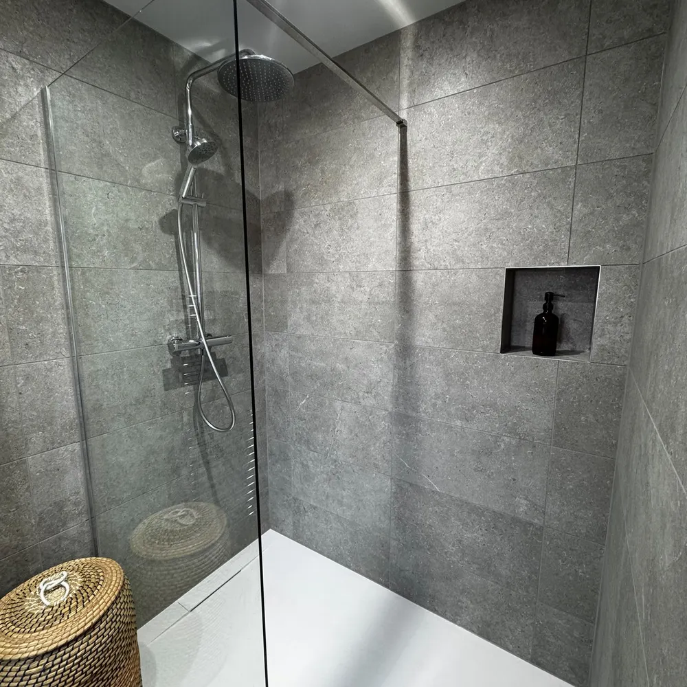 Level-access shower with a glass screen, a white tray set flush with the floor, a rain shower head and a recessed niche in grey stone-effect tile