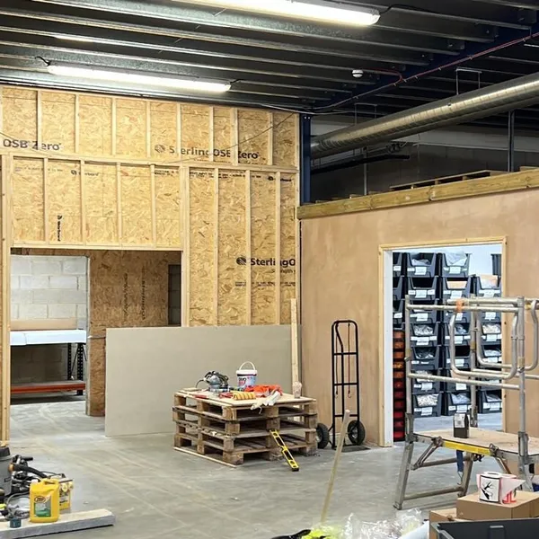 OSB sheathed stud walls beside a newly lined room whose doorway opens onto racks of stock bins, with a mobile scaffold tower in front