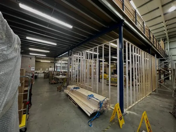 Timber stud framing set out between the steel columns under the mezzanine, with the warehouse still working around it